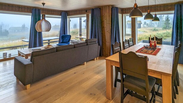 A spacious living area with a wooden ceiling, a large window with a view of a forest, a dining table with chairs, and a blue sofa.