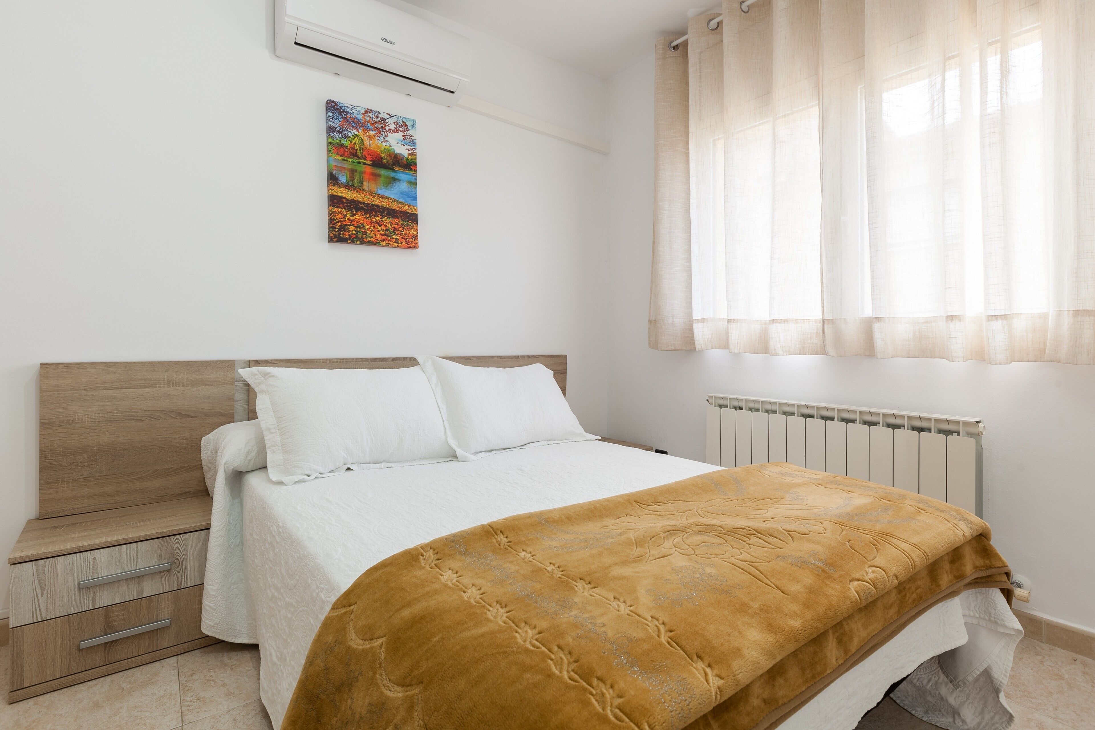 Un dormitorio con cama, mesita de noche, radiador y ventana con cortinas.