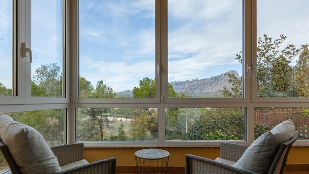 A room with two wicker chairs, a small round table, and large windows offering a view of trees and mountains.