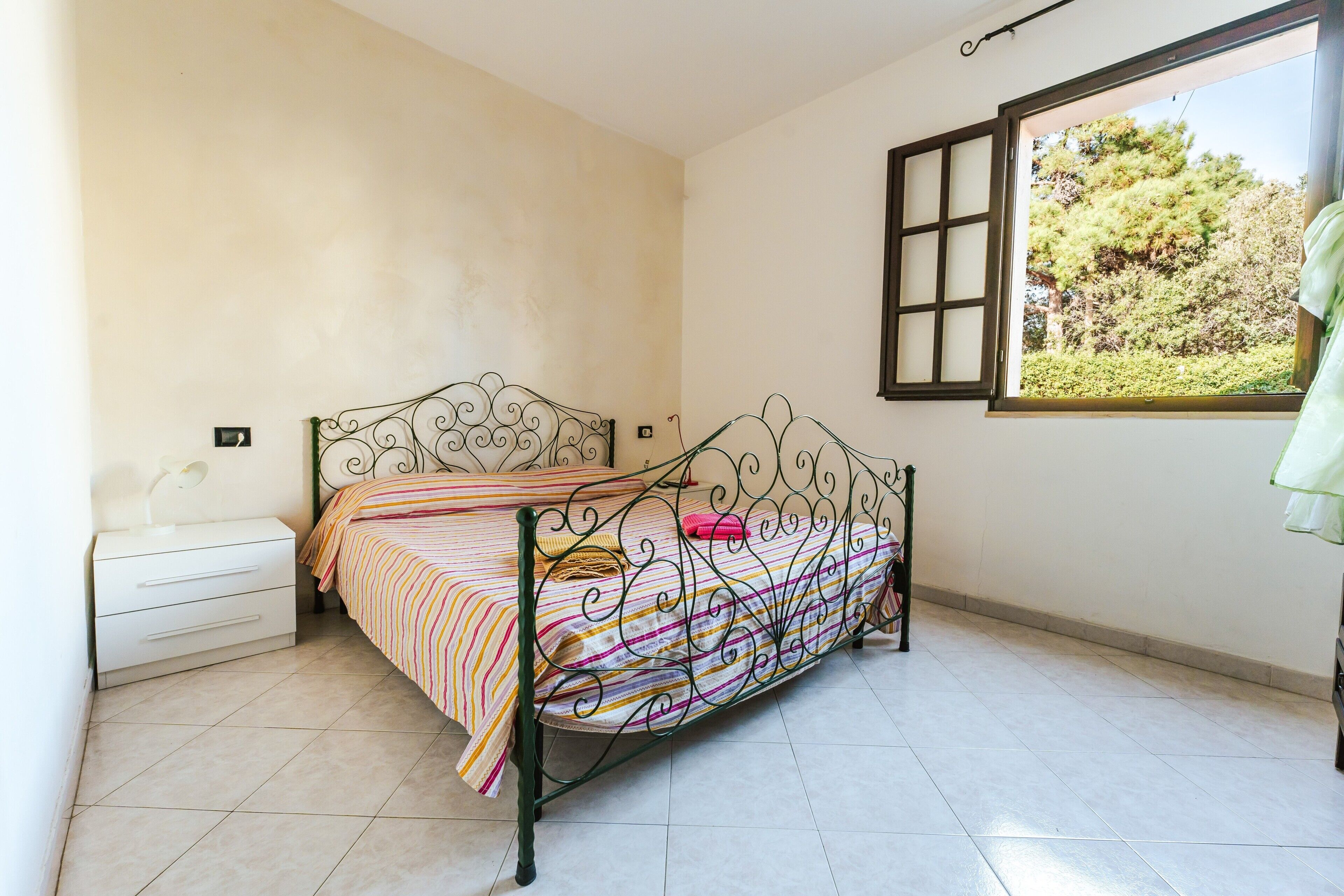 A single bed with a metal frame, a bedside table, and a window with a view of trees.