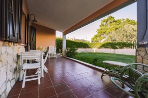 A covered patio with tiled flooring, outdoor furniture, and a view of a well-maintained garden and trees.