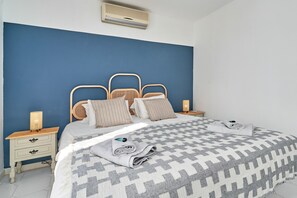 A bedroom with a blue wall, a bed with a patterned bedspread, two bedside tables with lamps, and a wall-mounted air conditioner.