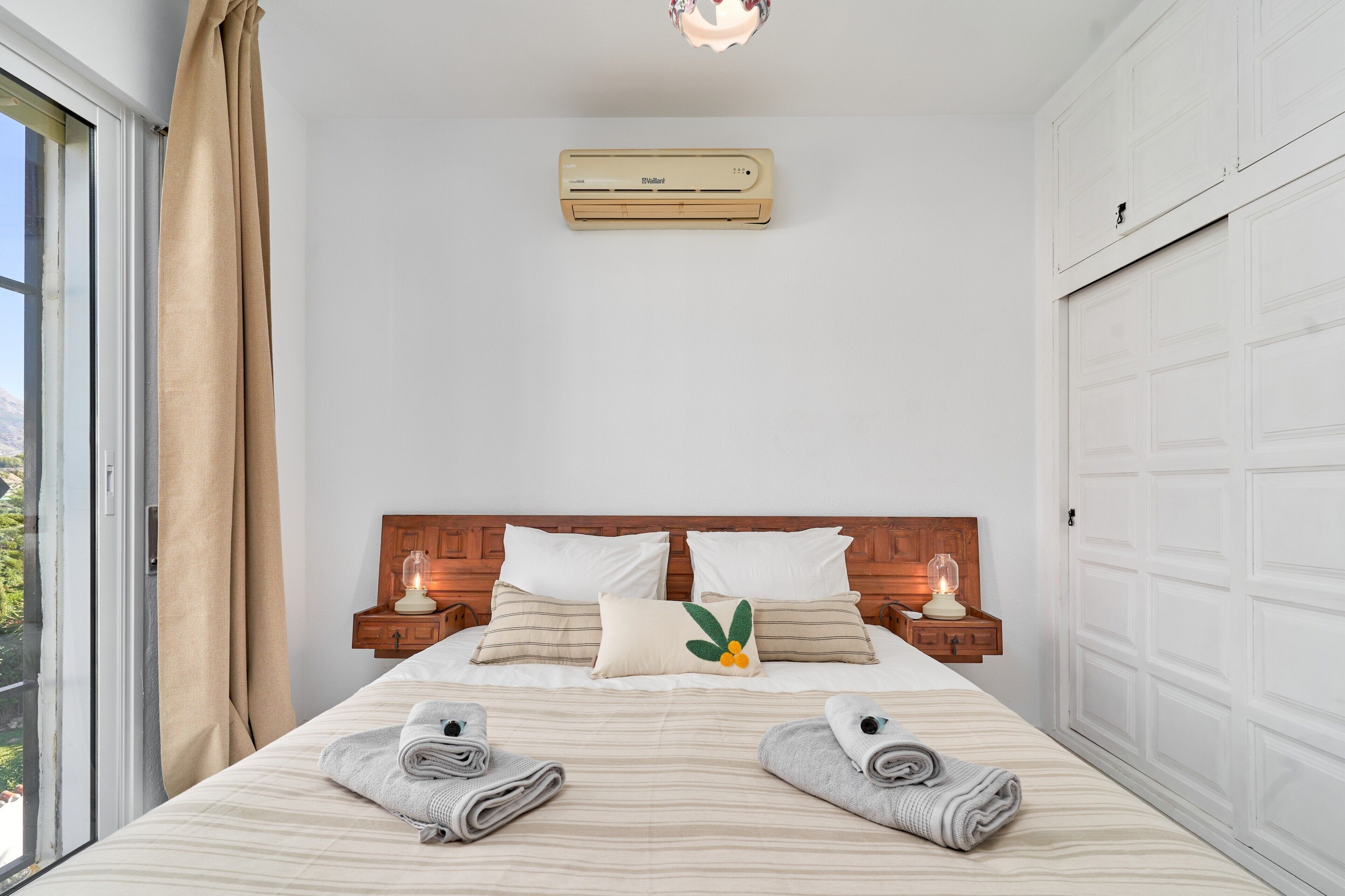 Habitación de hotel con cama, dos mesitas de noche, ventana con cortinas, equipo de aire acondicionado y vistas a zonas verdes.