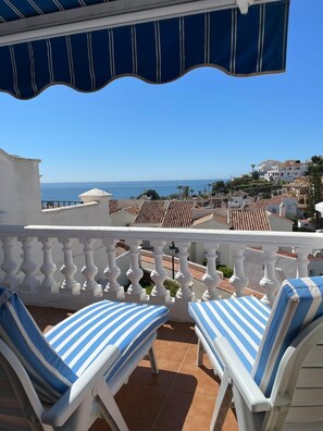 A balcony with white balustrades, two striped lounge chairs, and a view of the sea and coastal buildings.