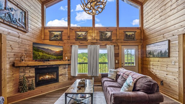 A log cabin interior with a fireplace, wooden walls, a large window, and a view of the outdoors.
