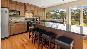 A modern kitchen with wooden cabinets, a large island with bar stools, a built-in oven, and a view of a lush garden through large windows. - Monthly Stays Available! Pool/WalkToBeach/1MileToSaltyDog/LagoonSetting/Relaxing (Hilton Head Island)