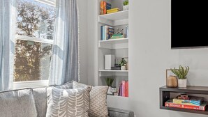 Un salon avec un canapé gris, des coussins à motifs, une bibliothèque remplie de livres et d’objets décoratifs, et une fenêtre avec des rideaux.