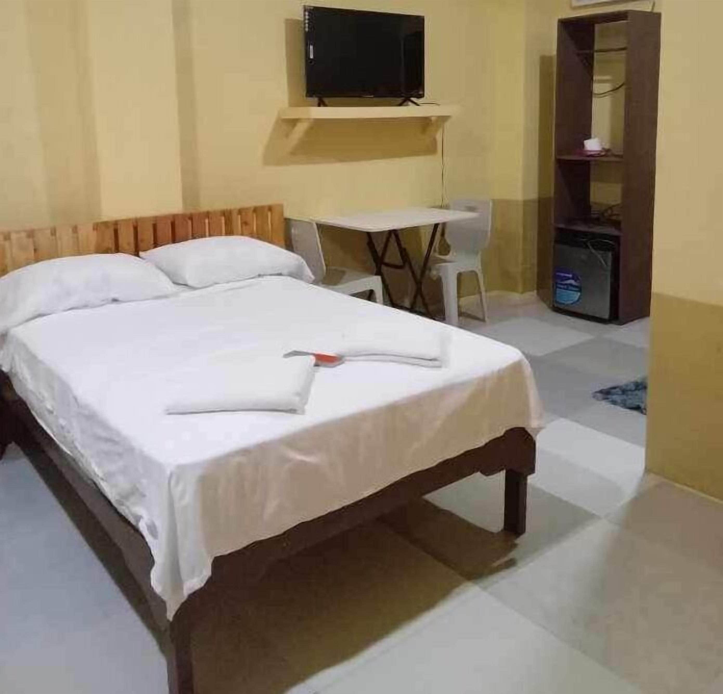 A neatly made bed with white linens, a small table with chairs, a television on a shelf, and a wardrobe.