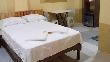 A neatly made bed with white linens, a small table with chairs, a television on a shelf, and a wardrobe.