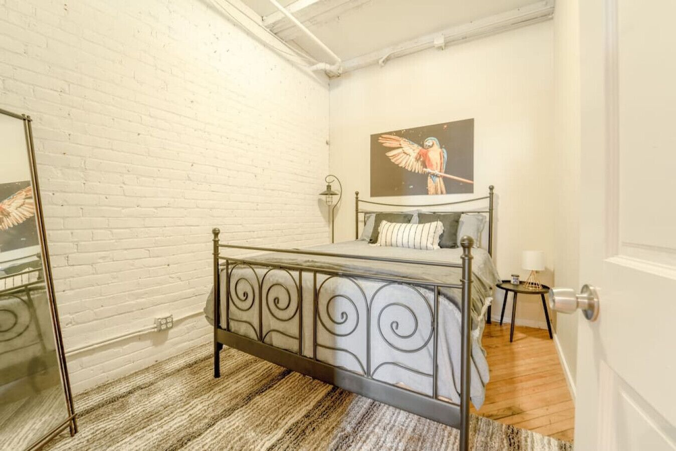 Une chambre avec un lit en métal, un mur en briques blanches, un plancher en bois, un tableau représentant un oiseau et une petite table avec une lampe.