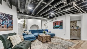 A modern living room with a sectional sofa, a coffee table, and a large rug. There is a painting of a car on the wall, and a kitchen area is visible through an open door. - Two Level Apt: Dtwn Buffalo (Buffalo)