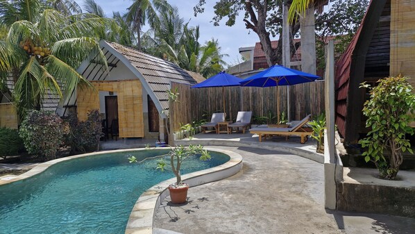 A pool area with lounge chairs and umbrellas, surrounded by tropical plants and a bamboo fence.