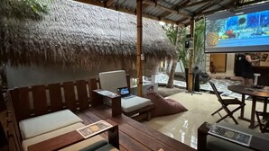 Un área de estar al aire libre con techo de paja, mobiliario de madera, una pantalla de televisión y una pequeña mesa con la carta.