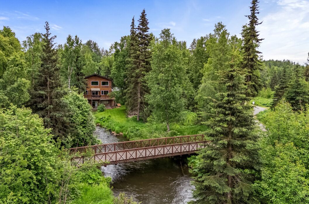 A lodge surrounded by dense forest, a wooden bridge over a stream, and a clear blue sky.