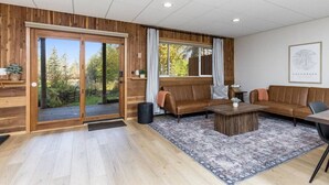 A spacious living room with a large sliding glass door leading to a deck, a brown leather sofa, a coffee table, and a rug. - Creekside Getaway: On the Creek in Anchorage (Anchorage)