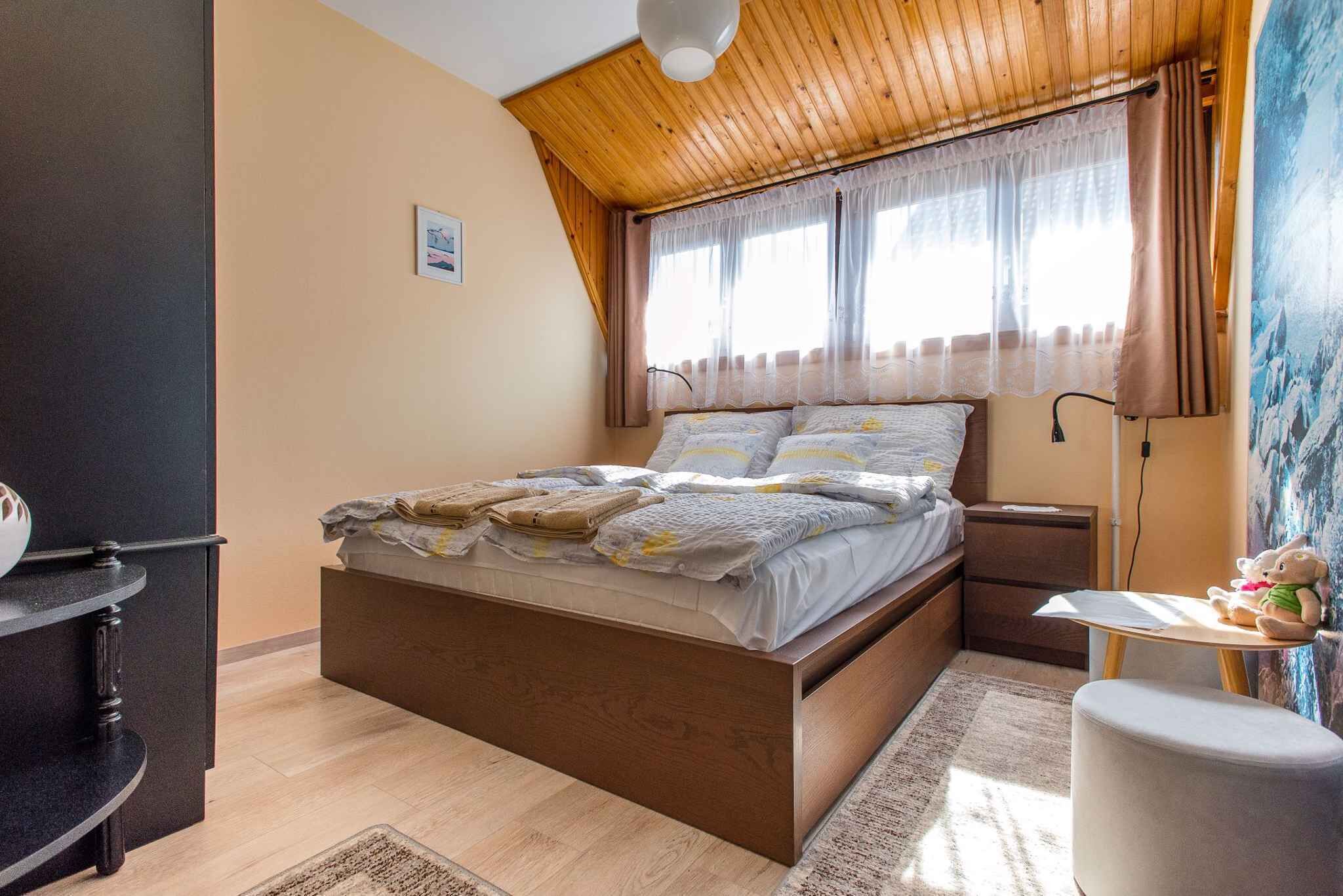 Une chambre avec un plafond en bois, un lit recouvert d’une couette blanche et grise, une table de chevet avec une lampe et un petit tapis.