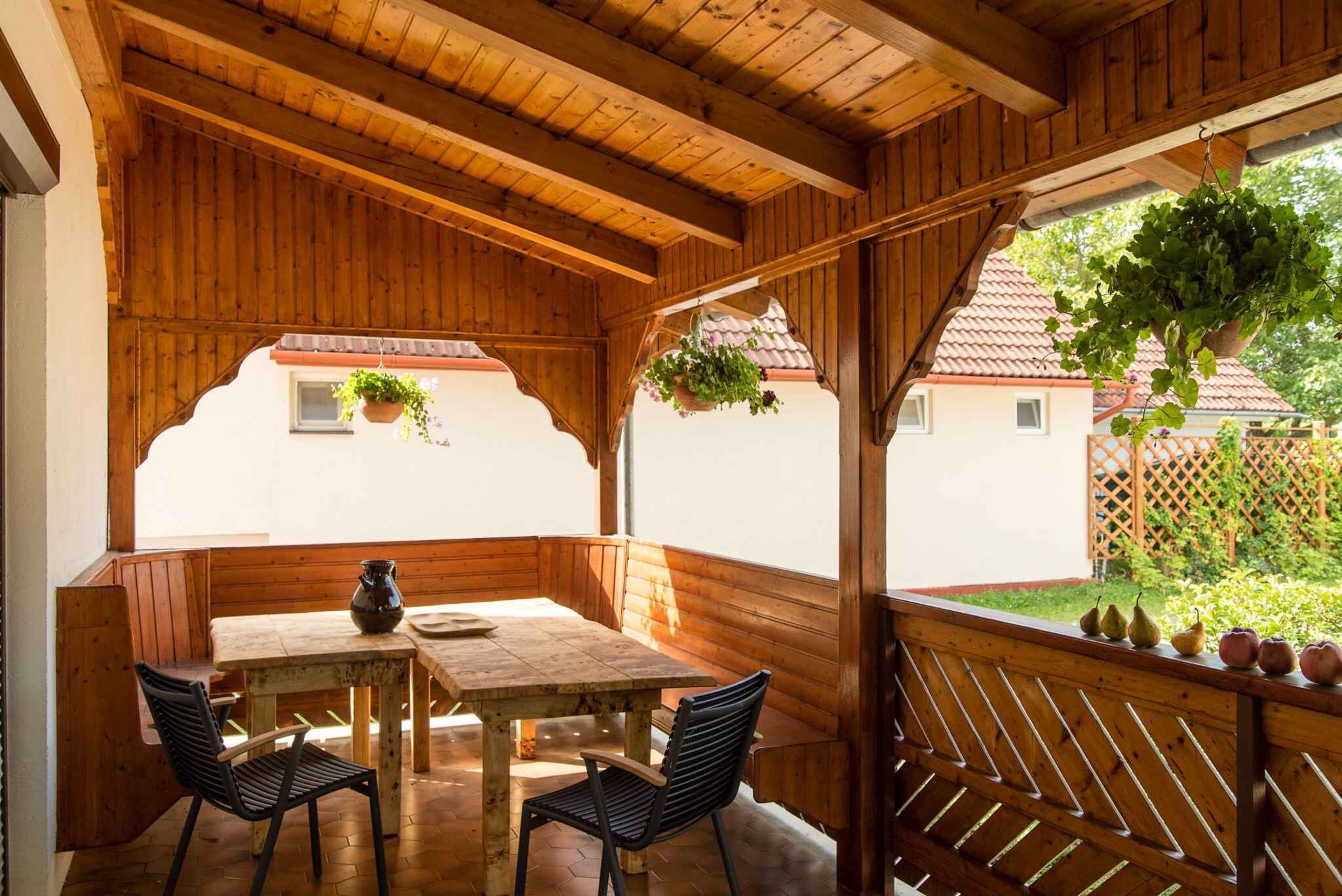 Ein Holz-Sitzbereich im Freien mit Tisch und Stühlen, einer hängenden Pflanze und Blick auf ein Haus mit roten Ziegeldach.