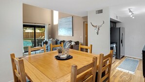 Zona pranzo con tavolo e sedie in legno, cucina con frigorifero e vista su un giardino con piscina.