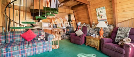 A cozy cabin interior with a plaid sofa, purple armchairs, a spiral staircase, a fireplace, and a rustic stone wall.