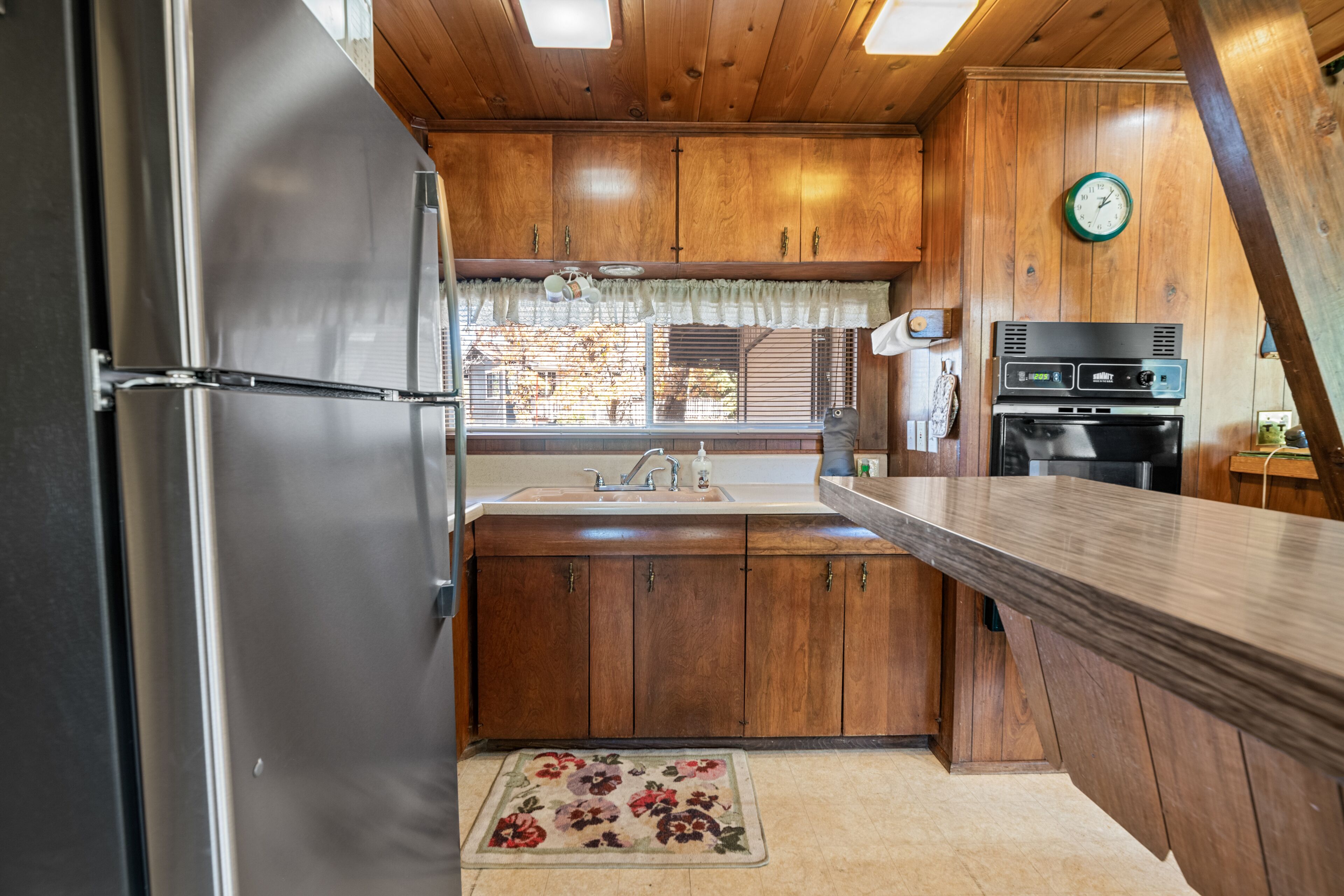 A kitchen with wooden cabinets, a stainless steel refrigerator, a sink, and a clock on the wall.