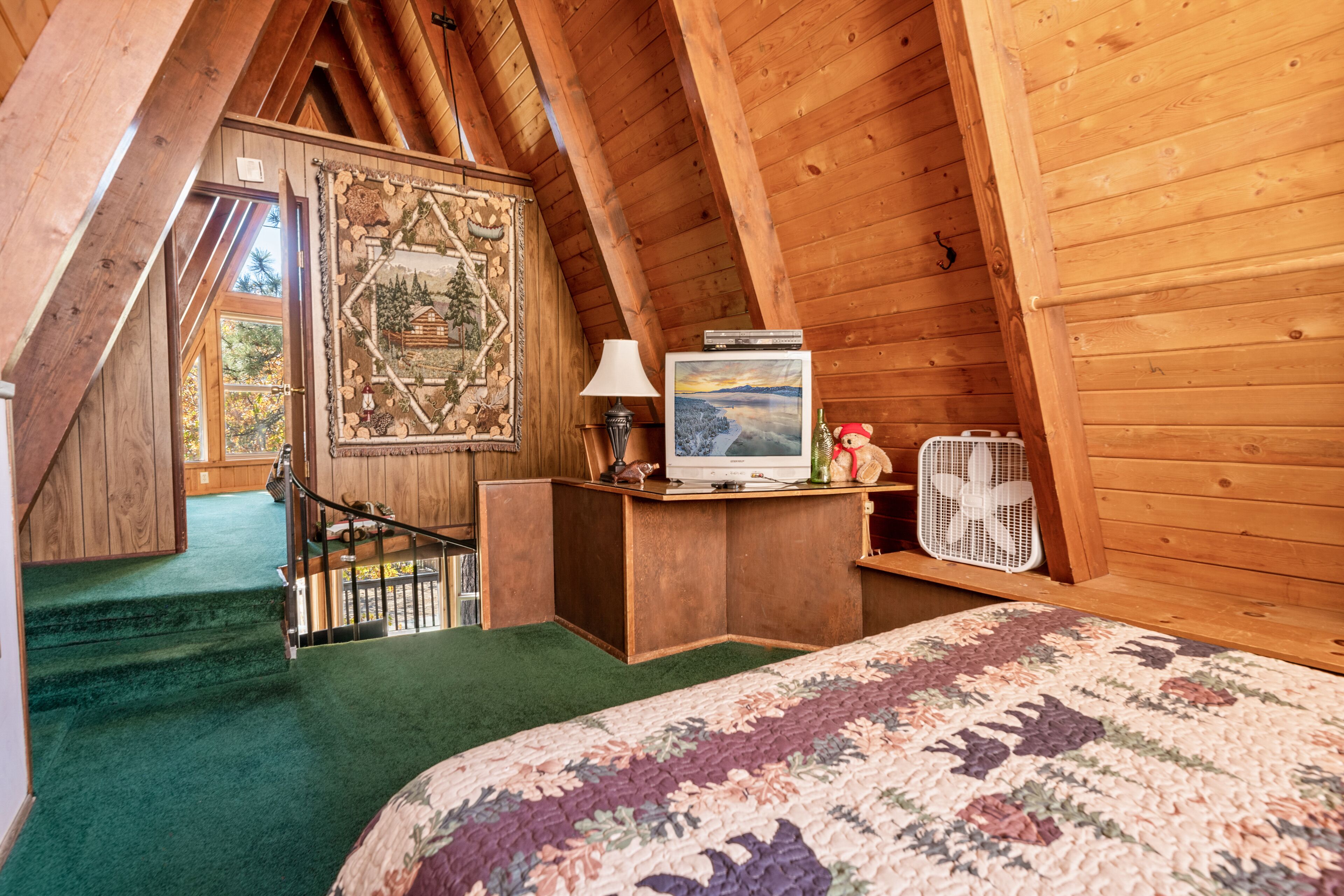 Un dormitorio cálido en el ático con techo de madera, una cama con colcha floral, un escritorio con lámpara y televisor, y un ventilador.