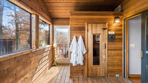 Una cabaña de madera con un porche, una puerta que da a un baño y una ventana con vistas a los árboles.