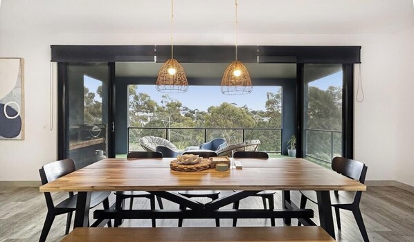 A dining area with a wooden table, black chairs, wicker hanging lights, and a view through large glass doors.