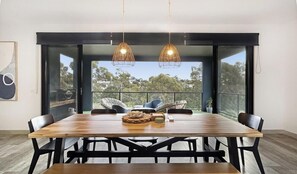 Un espace repas avec une table en bois, des chaises noires, des suspensions en osier et une vue dégagée grâce à de grandes portes vitrées.