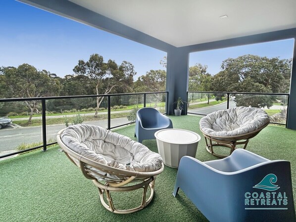 A balcony with wicker chairs, a round table, and a view of the road and trees.