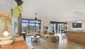 A modern living room with a sofa, coffee table, and a dining area with chairs. There are large windows providing natural light and a view of the outdoors.