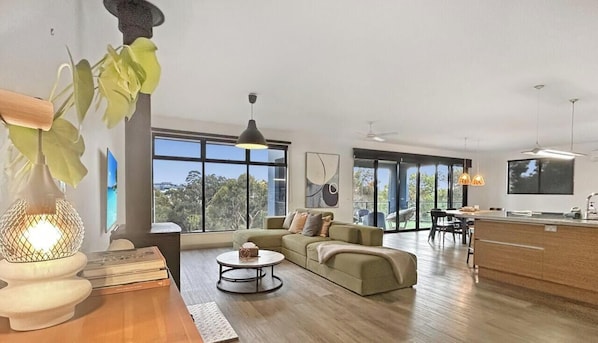 Un salon moderne comprenant un canapé, une table basse et un coin repas avec des chaises. De grandes fenêtres laissent entrer la lumière naturelle et offrent une vue sur l’extérieur.