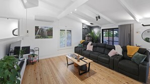 A modern living room with a sectional sofa, a coffee table, and a TV. There are windows with blinds, a fireplace, and a bookshelf.