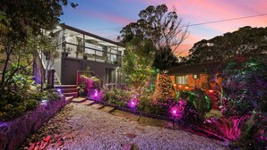 A modern house with a balcony, surrounded by a garden with colorful lights and a gravel pathway.