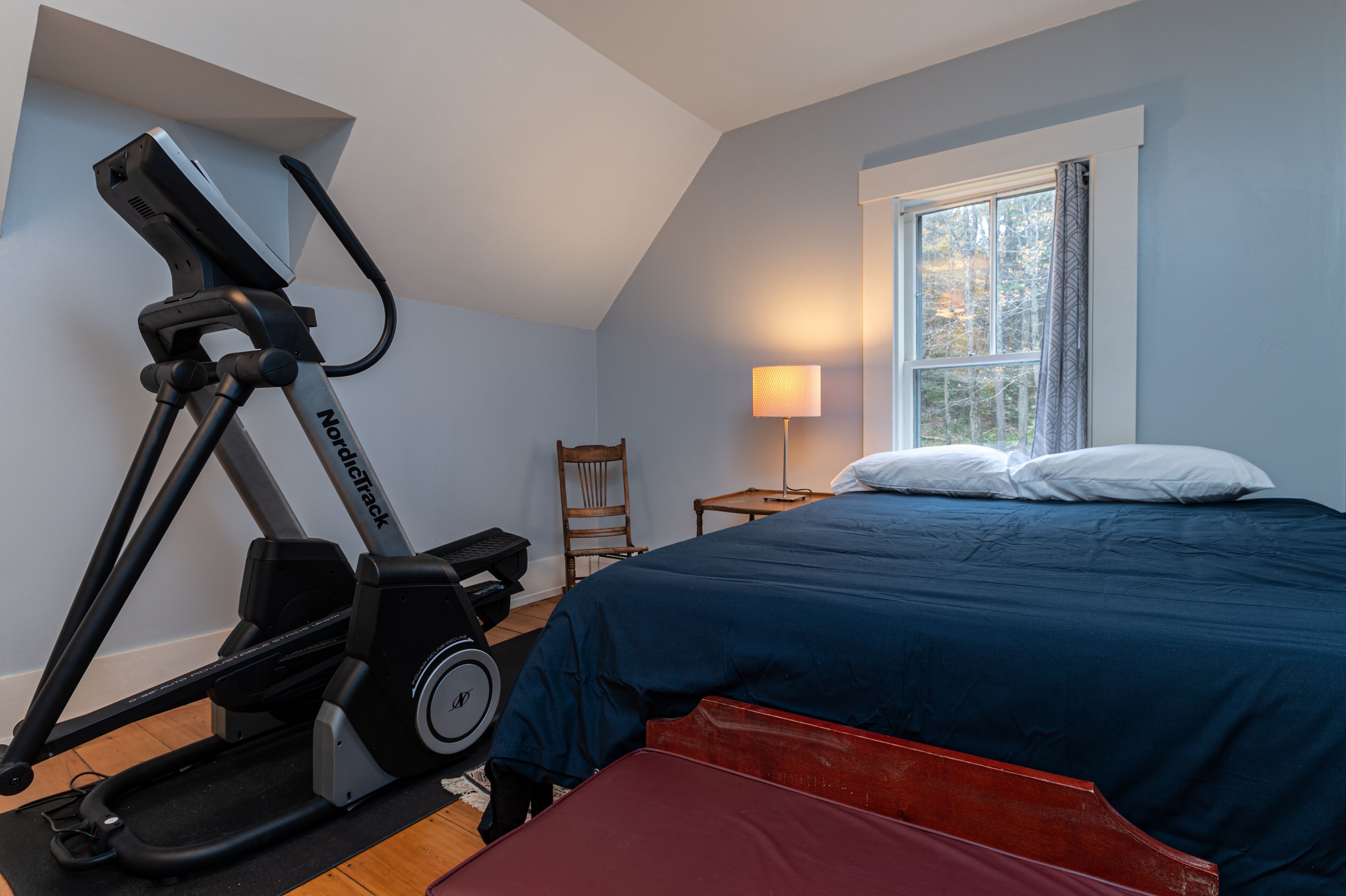 A bedroom with a bed, a standing exercise bike, a chair, a lamp, and a window.