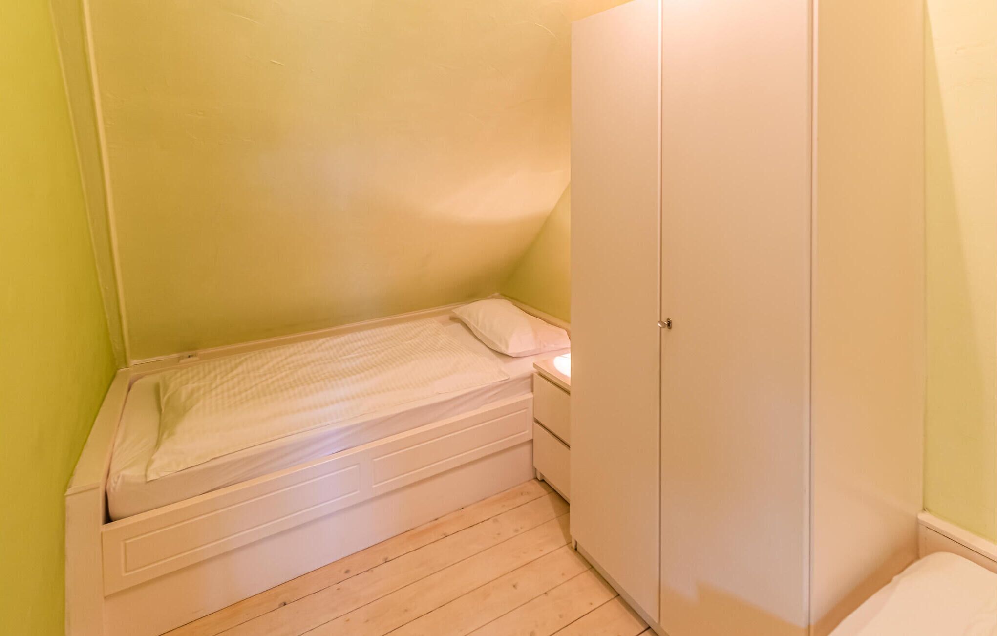 Une petite chambre avec un lit simple, un plancher en bois, une armoire blanche et une table de chevet.