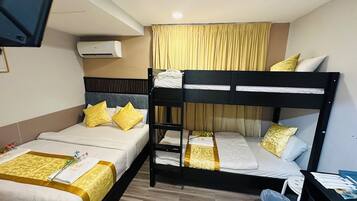 A hotel room with two beds, one with a bunk, a TV, an air conditioner, and a small trash bin.