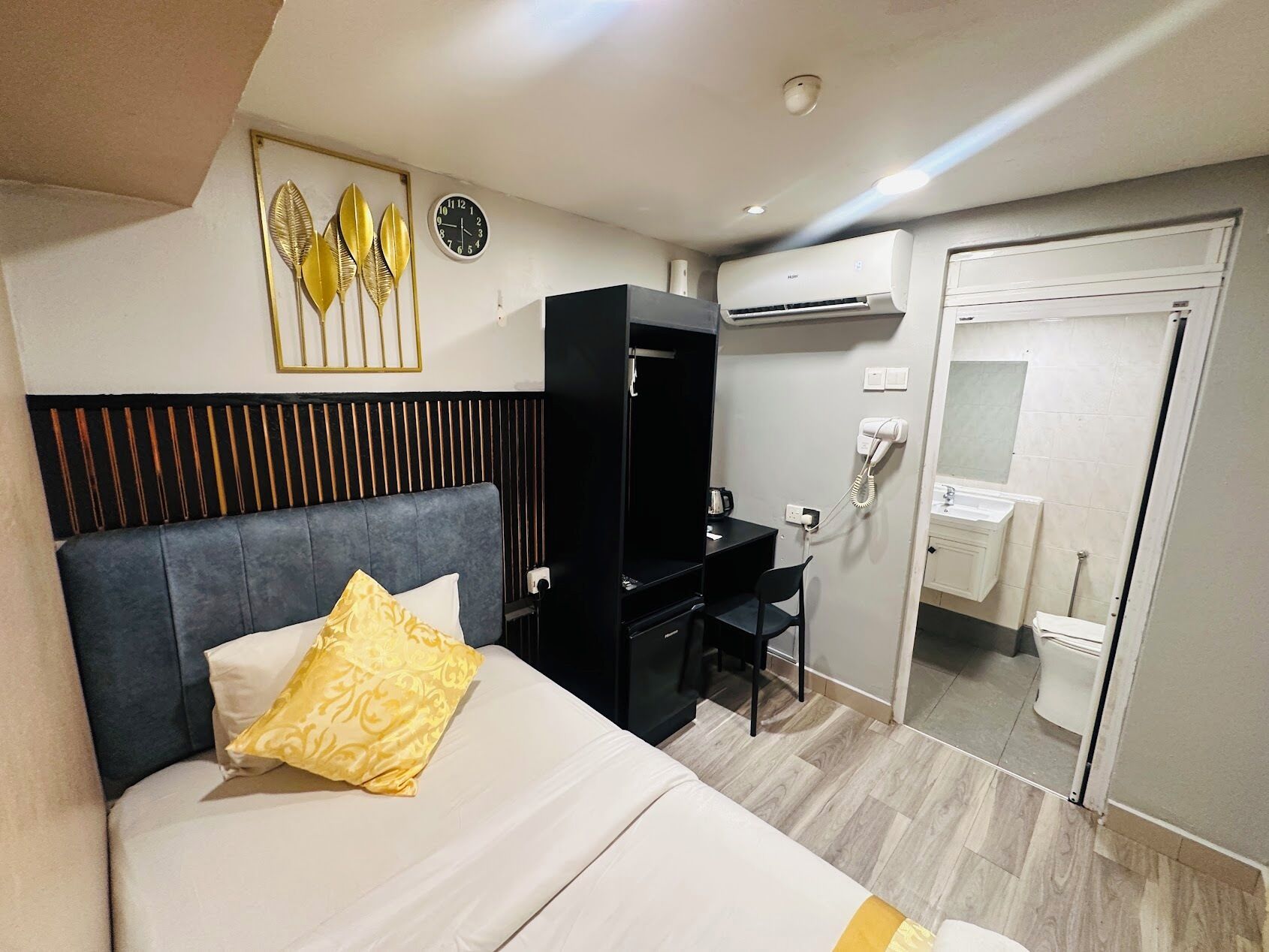 A hotel room with a bed, a desk, a chair, an air conditioning unit, a telephone, and a bathroom visible through an open door.
