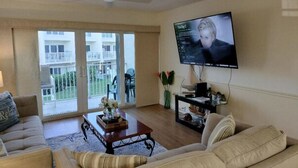 A living room with a television mounted on the wall, a sofa, a coffee table, and a sliding glass door leading to an outdoor area. - Center of Saint Augustine Beach (St. Augustine)