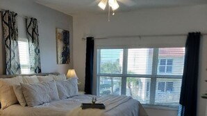 A bedroom with a large bed, a ceiling fan, and a window with a view of buildings. - Center of Saint Augustine Beach (St. Augustine)
