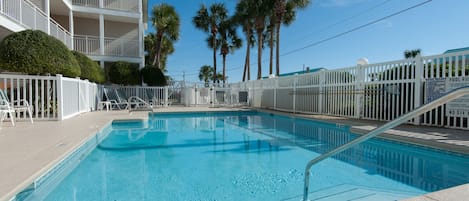 A swimming pool with a white fence, palm trees, and a multi-story building in the background.