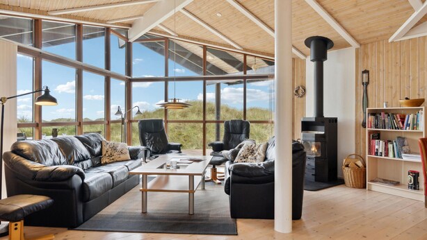 A spacious living room with a large window, a black leather sofa, a coffee table, and a bookshelf. - Beautiful home in Saltum (Saltum)