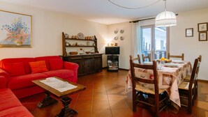 A living room with a red sofa, a dining table with chairs, a kitchen area with shelves, and a large window with a view. - Lovely apartment for 5 guests with WIFI, TV and balcony (Canazei)