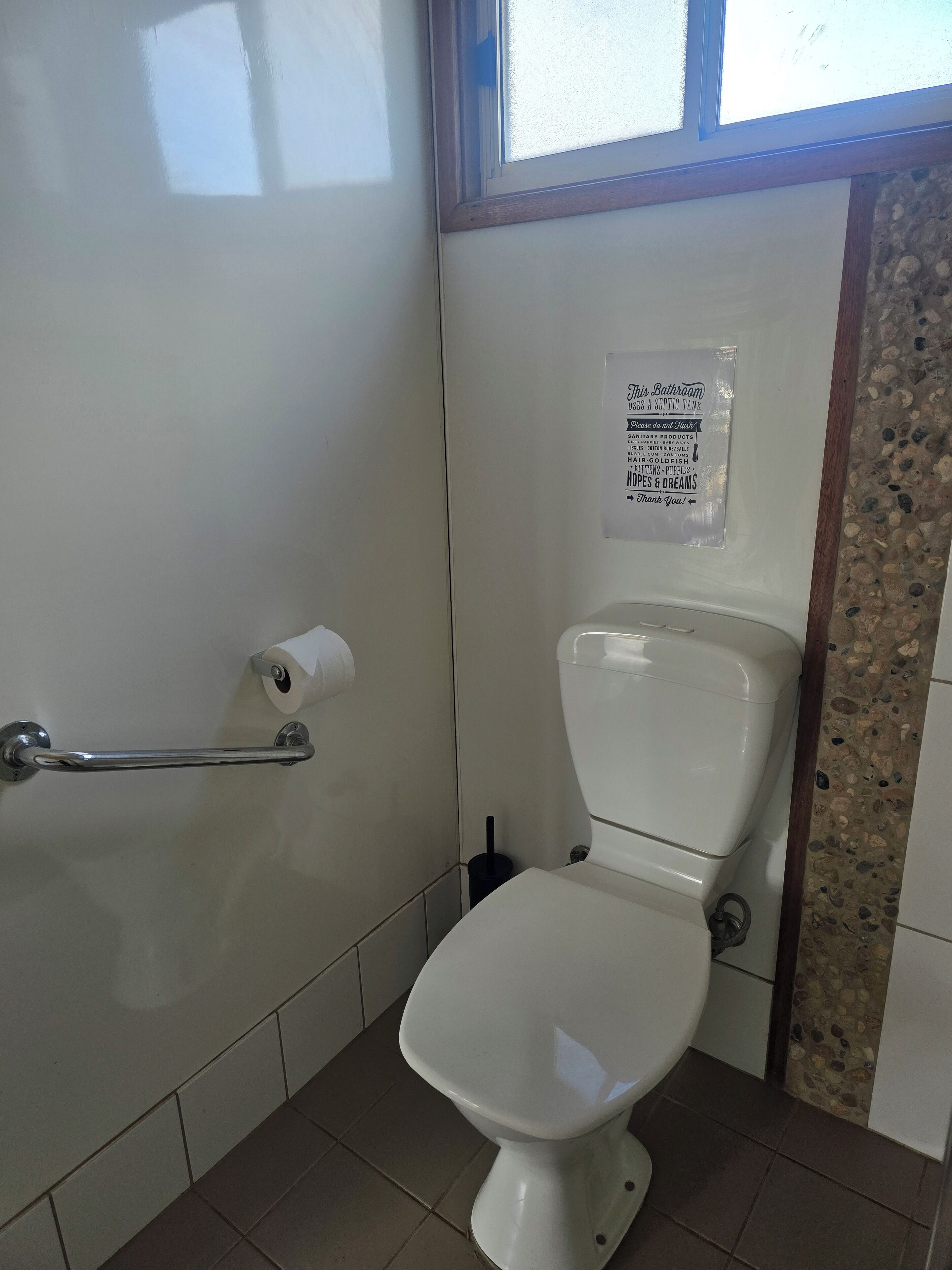 A bathroom with a white toilet, a handrail, a paper towel dispenser, and a sign indicating a septic tank.