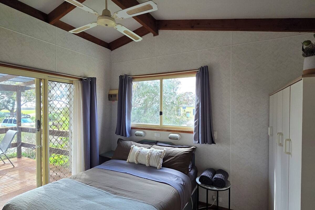 Une chambre à coucher comprenant un lit, une petite table, une armoire, un ventilateur de plafond et une fenêtre avec des rideaux.