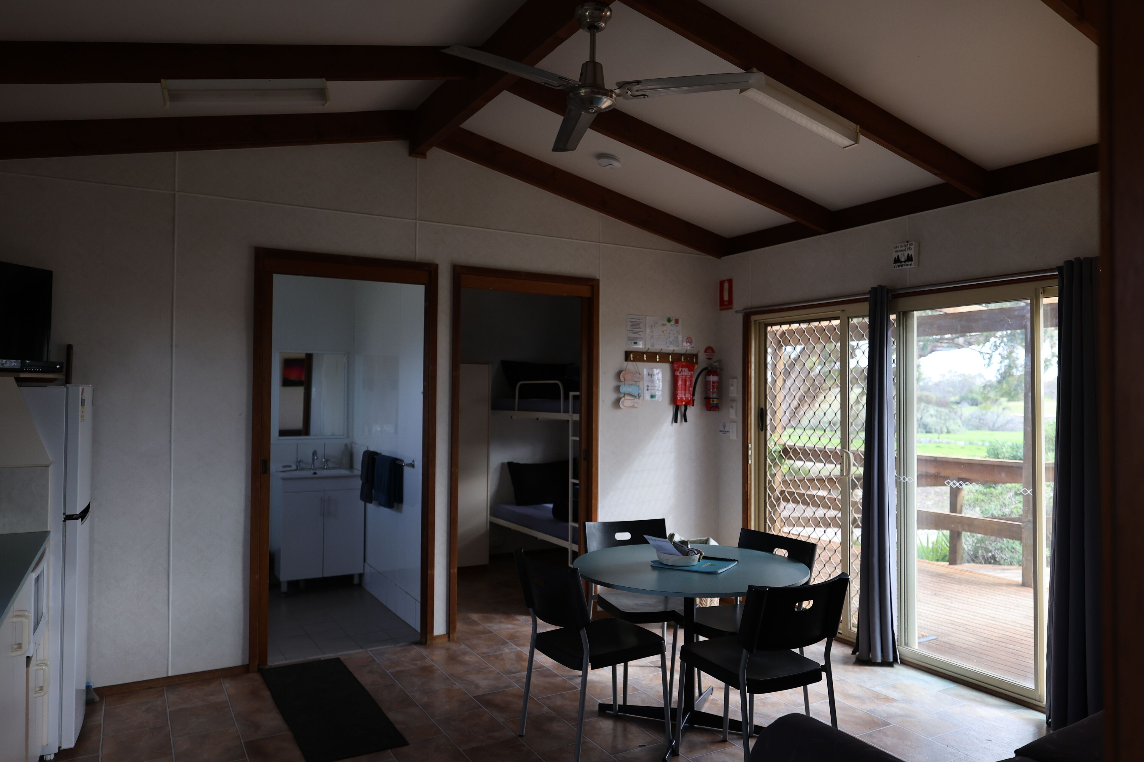 A room with a dining table, chairs, a ceiling fan, a kitchen area, and a sliding glass door to the outside.