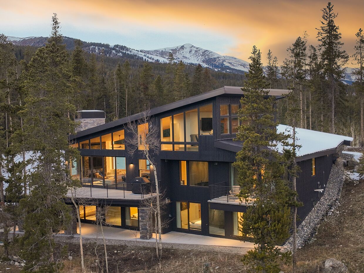 A modern two-story house with large windows, a deck, and a mountainous backdrop.