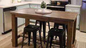 Een keuken met lichtblauwe kastjes, een houten tafel met twee krukken, een RVS magnetron en een vaatwasser.