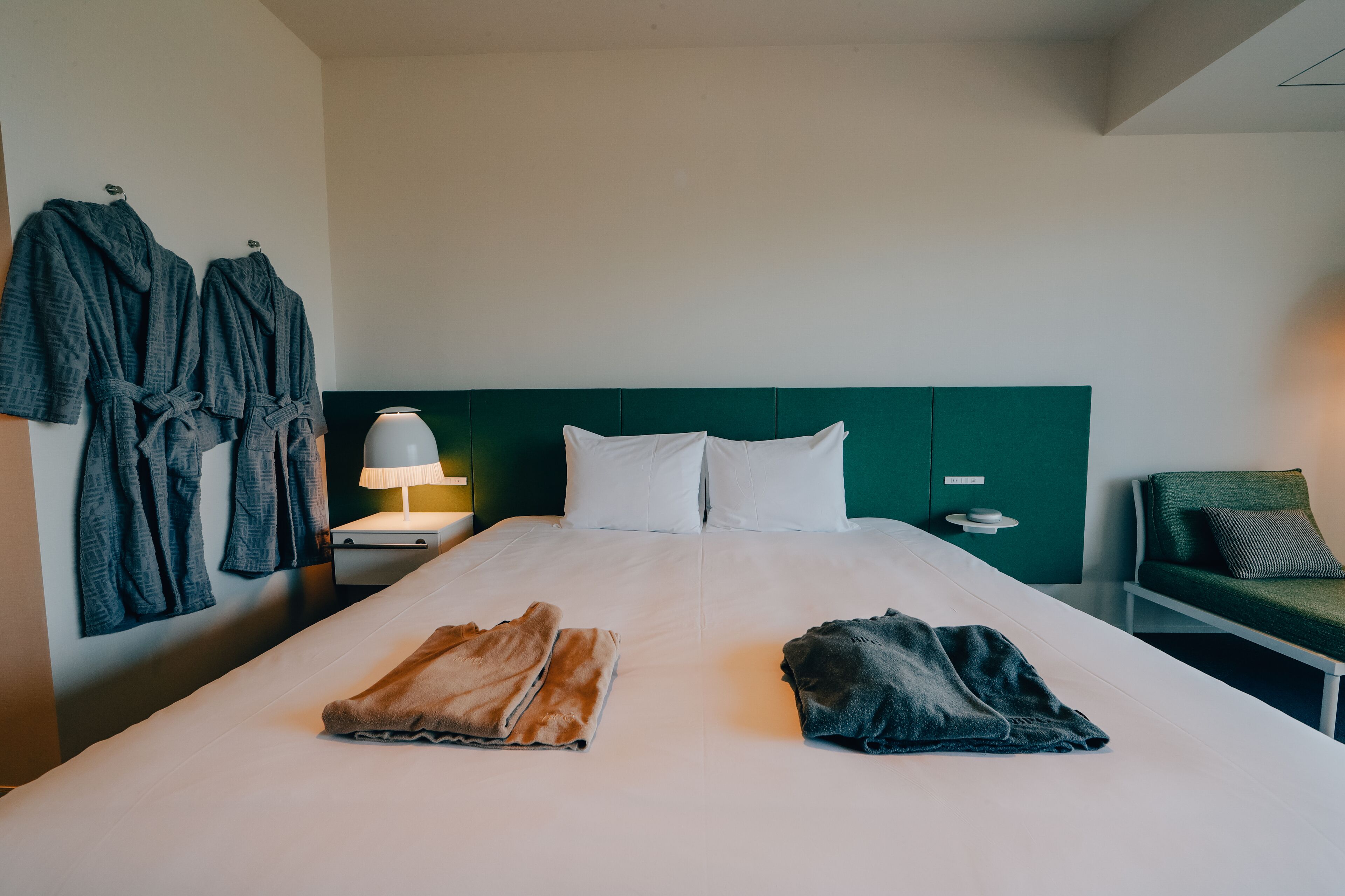A hotel room with a neatly made bed, two robes on the wall, a bedside table with a lamp, and a green chair.