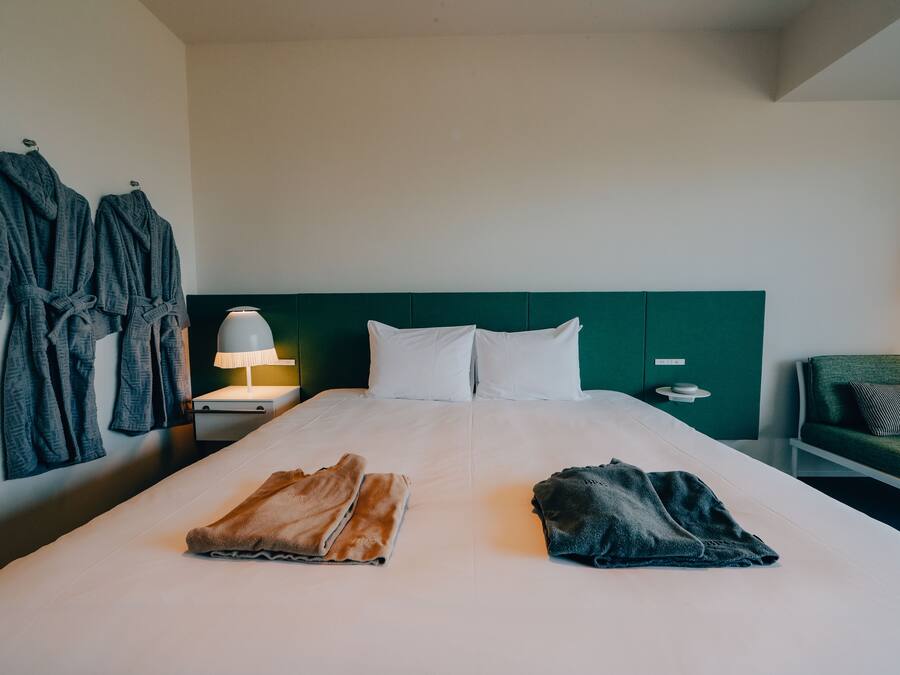 Ein Hotelzimmer mit einem ordentlich bezogenen Bett, zwei Bademänteln an der Wand, einem Nachttisch mit Lampe und einem grünen Sessel.