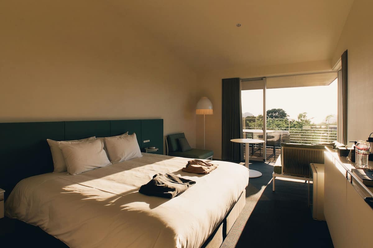 A hotel room with a large bed, a desk with a lamp, a chair, a small table, and a view of the outdoors.